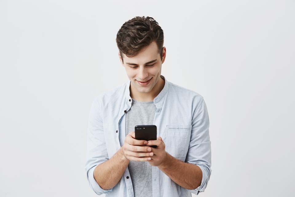 handsome-joyful-student-with-dark-hair-wearing-blue-shirt-messaging-typing-message-using-free-onlipe-app-his-smartphone-looking-screen-with-smile-posing